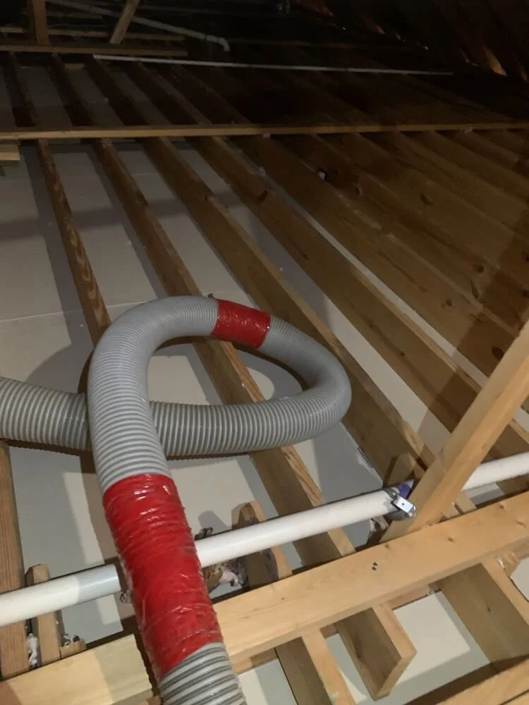 Exposed attic floor framing with visible wooden beams, pipes, and flexible ventilation duct.