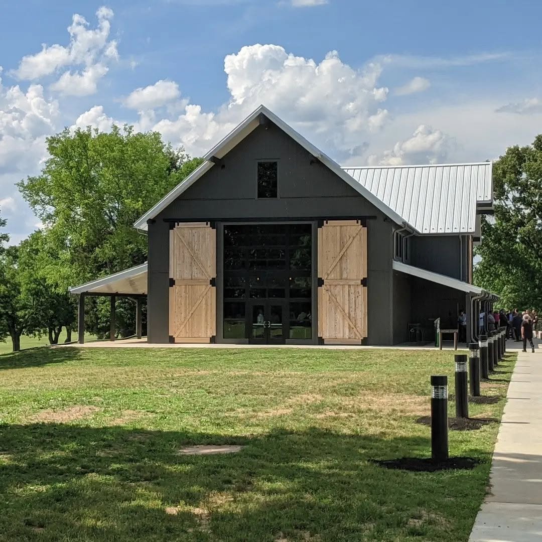 A modern barn with black exterior walls and light wood barn doors, situated on a grassy area with trees and a partly cloudy sky in the background.