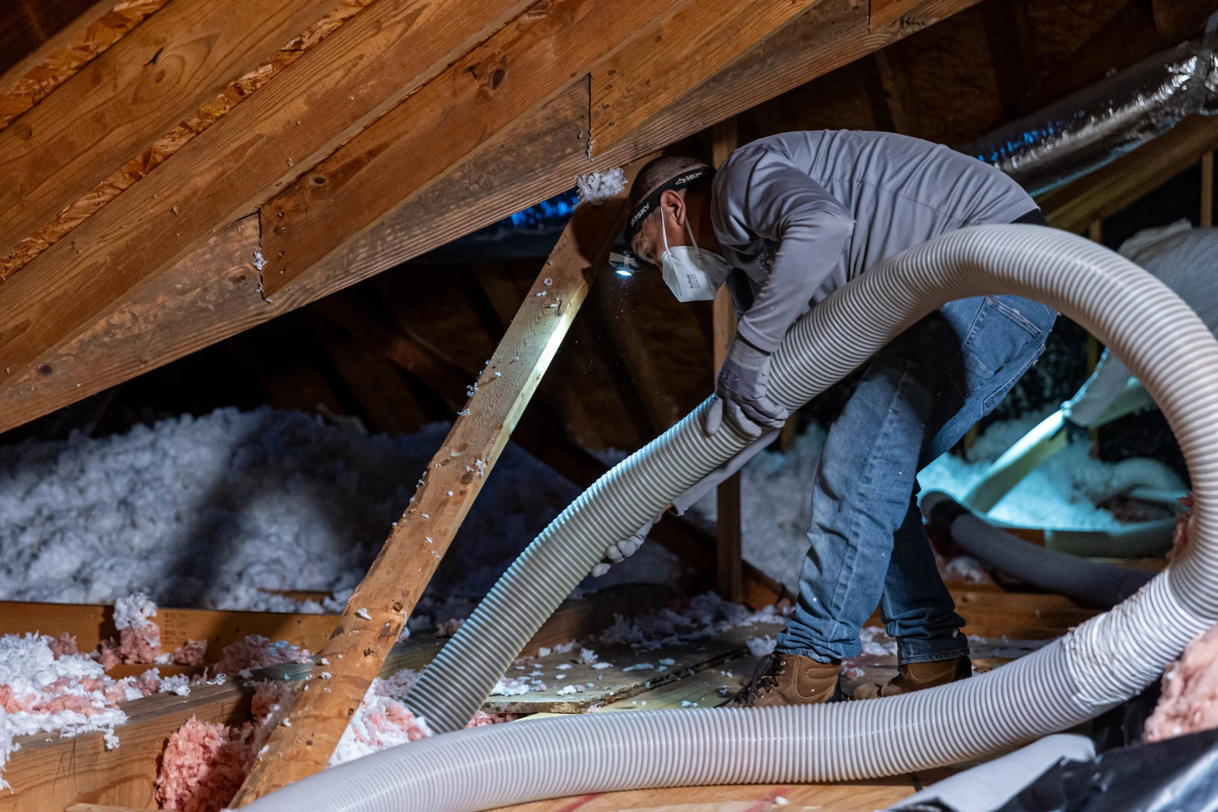 Worker wearing protective gear spraying foam insulation on attic ceiling.