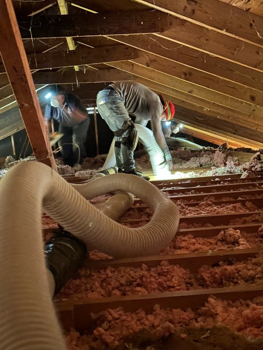 Two workers installing insulation in an attic, using a large flexible hose connected to insulation blowing equipment, with wood framing and a sloped ceiling.