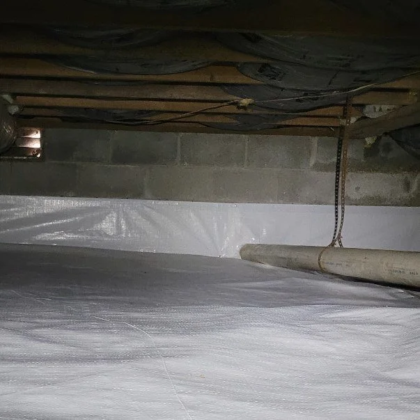 Basement with plastic sheeting on the floor, a concrete block wall, and a pipe along the floor.