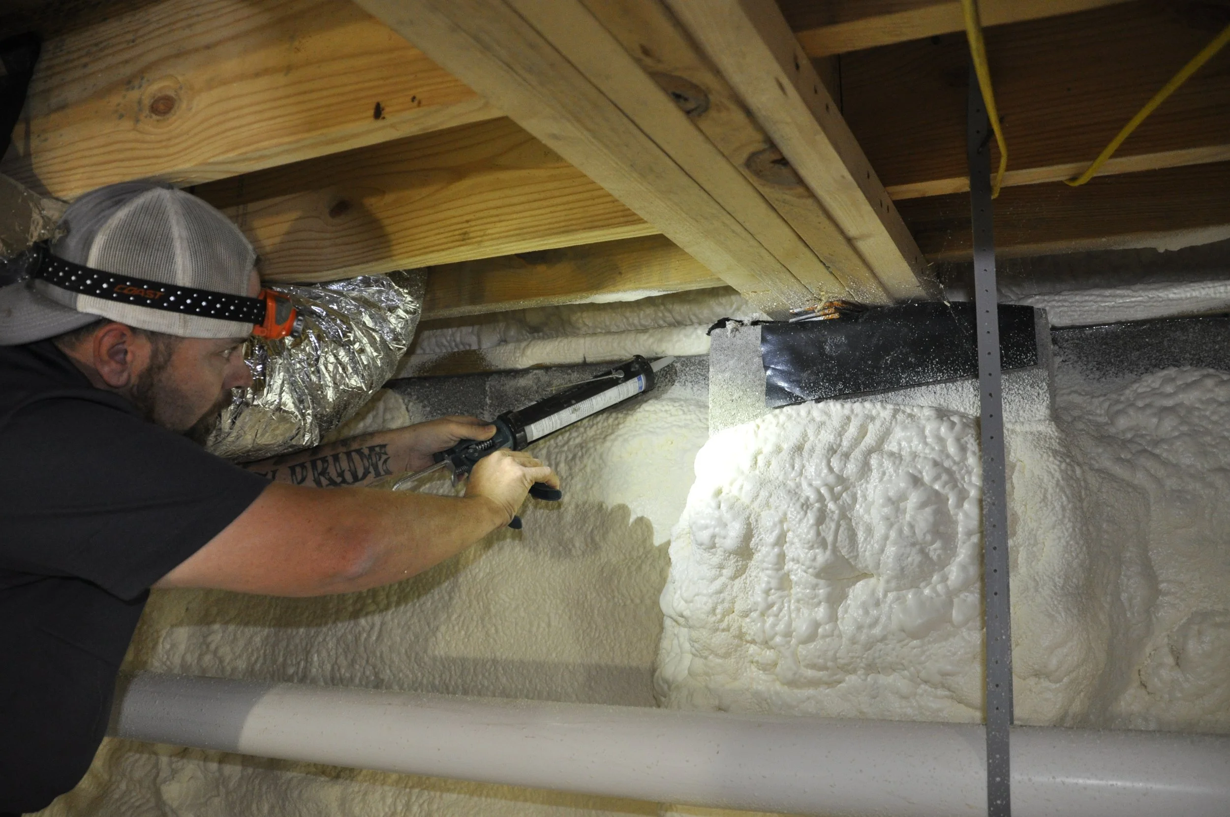 A person wearing a headlamp is spray foaming insulation into a wall cavity under a wooden ceiling.
