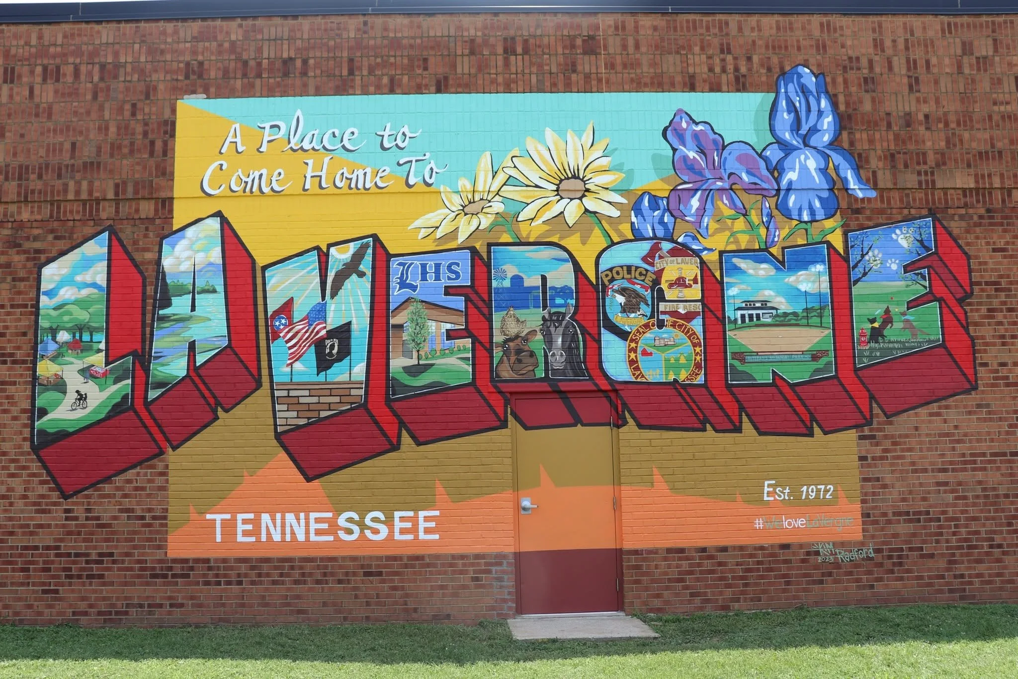 Street art mural on a brick wall that says 'A Place to Come Home To' above the word 'Tennessee' in large letters. The mural features colorful flowers, the Tennessee state flag, and city emblems.