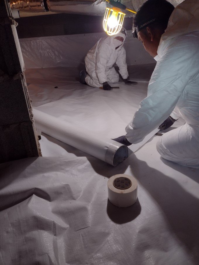 Two workers in protective suits installing or repairing a large white plastic or fabric sheet, working under a lamp at night, with rolls of tape nearby.