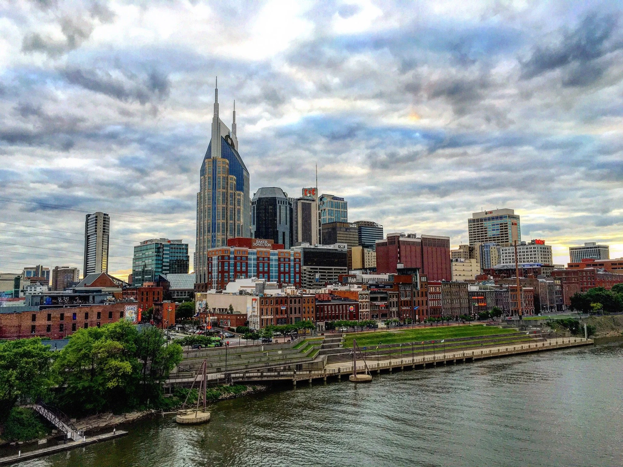 City skyline with numerous tall buildings under a cloudy sky, a river flowing in the foreground, and greenery along the riverbank.