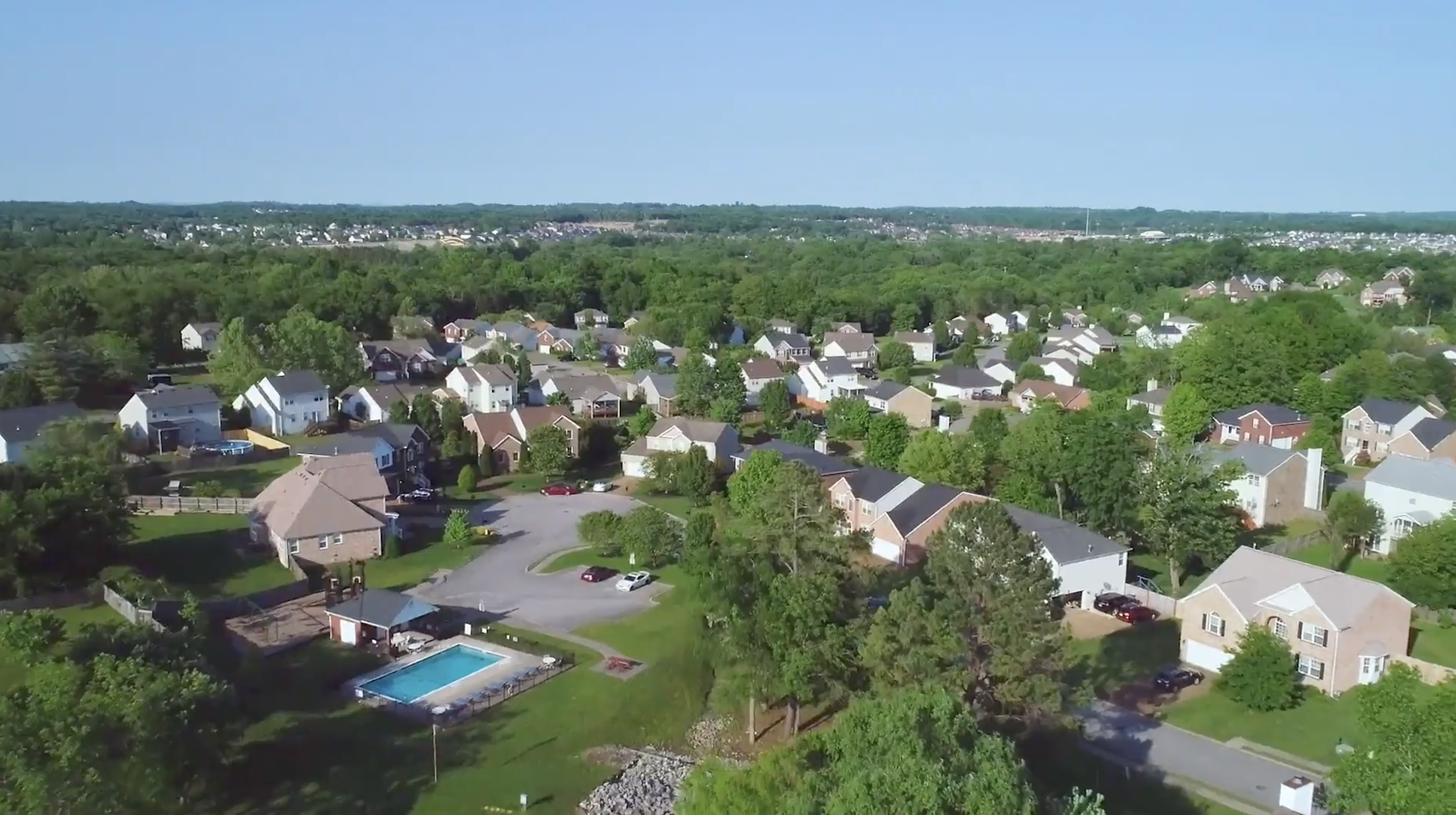 Aerial view of a suburban neighborhood with houses, green trees, a swimming pool, and a distant forest under a clear blue sky.