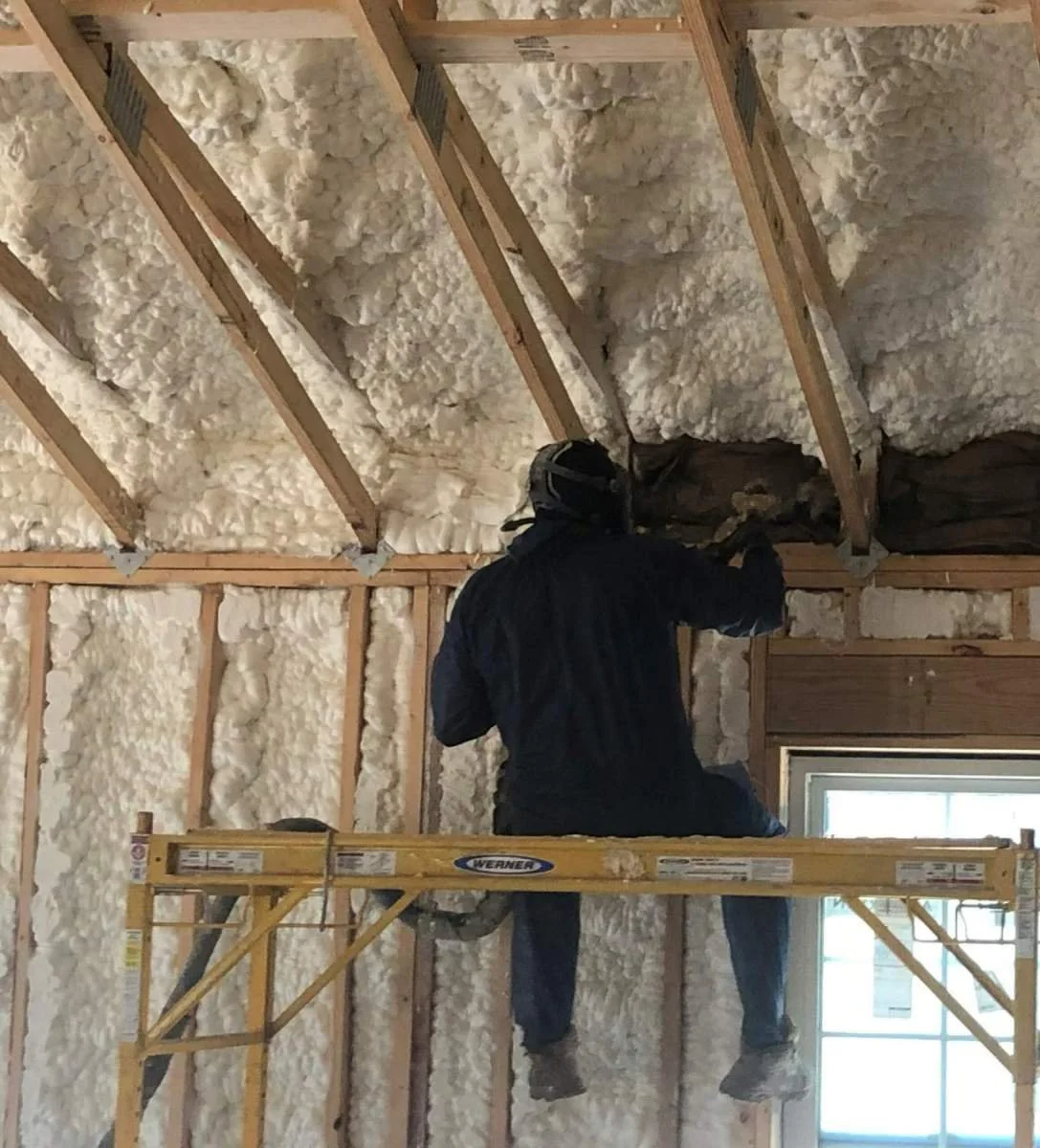 A worker on a yellow scaffolding installing insulation in an attic or wall cavity.