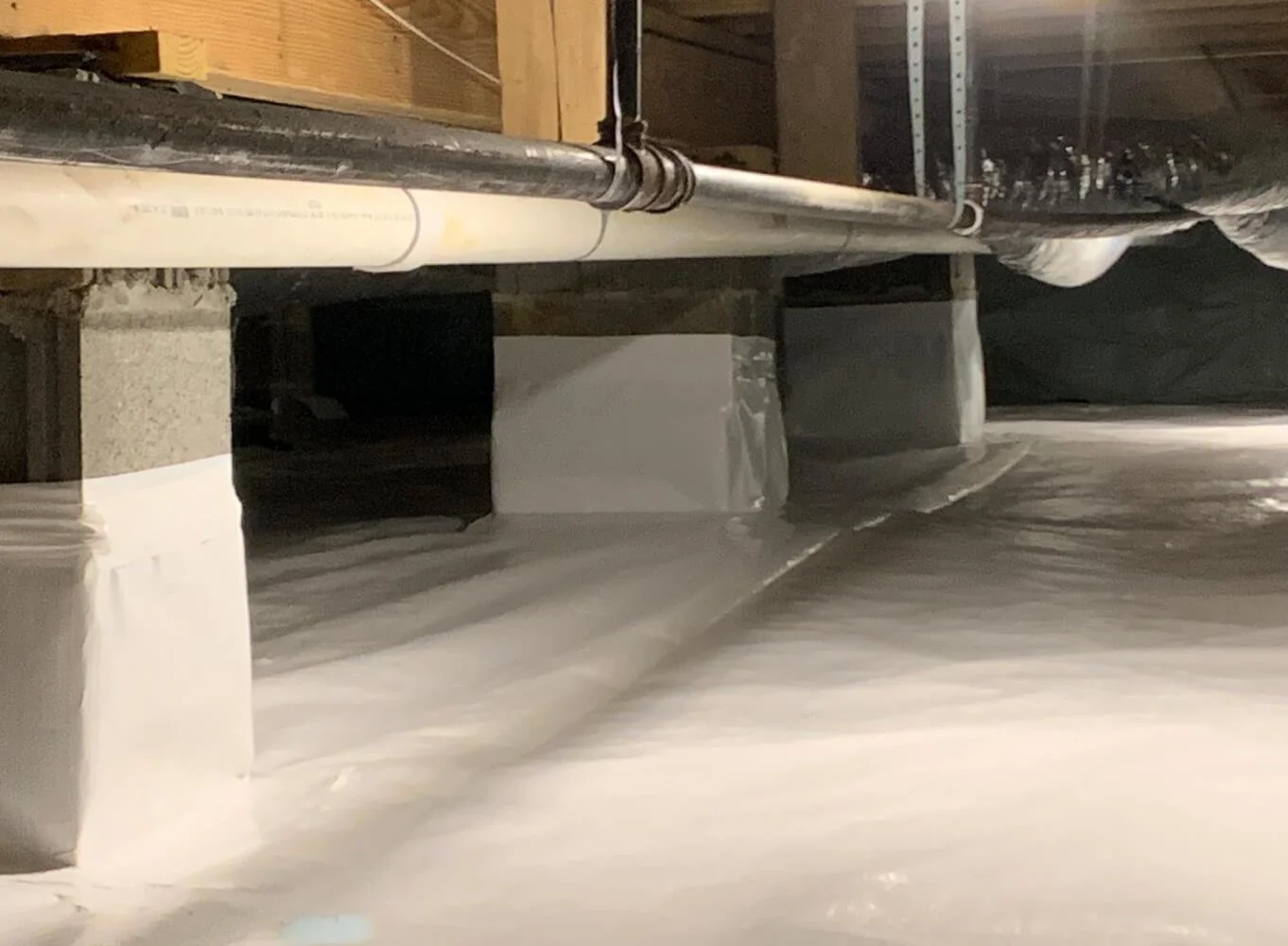 Underground foundation with concrete blocks supporting wooden framing, drainage pipes, and a concrete floor.