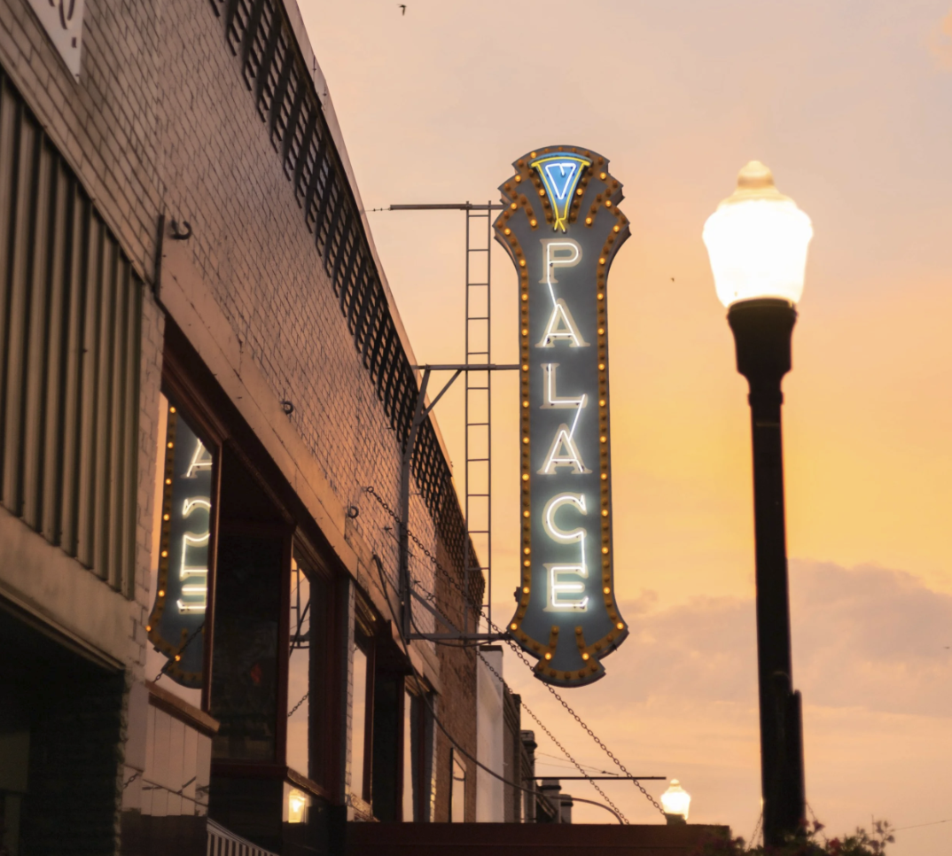Neon sign reading 'Palace' hanging on the side of a brick building during sunset, with street lamps illuminating the street.