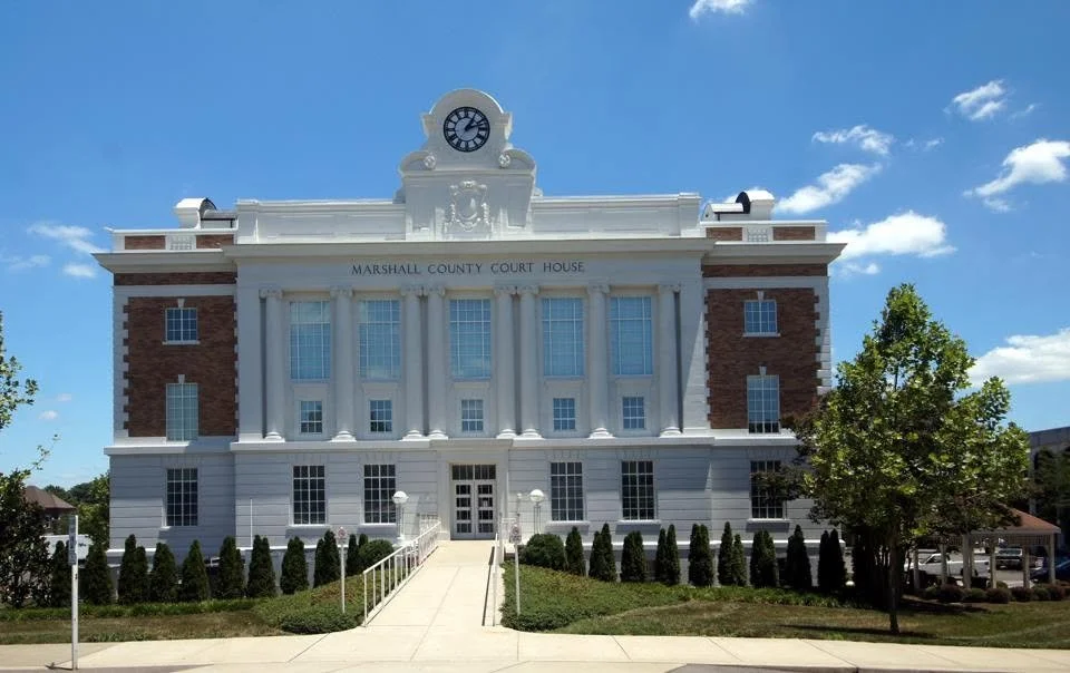 Front view of the Marshall County Courthouse, a historic building with a clock tower, white facade, and red brick accents, surrounded by greenery on a sunny day.