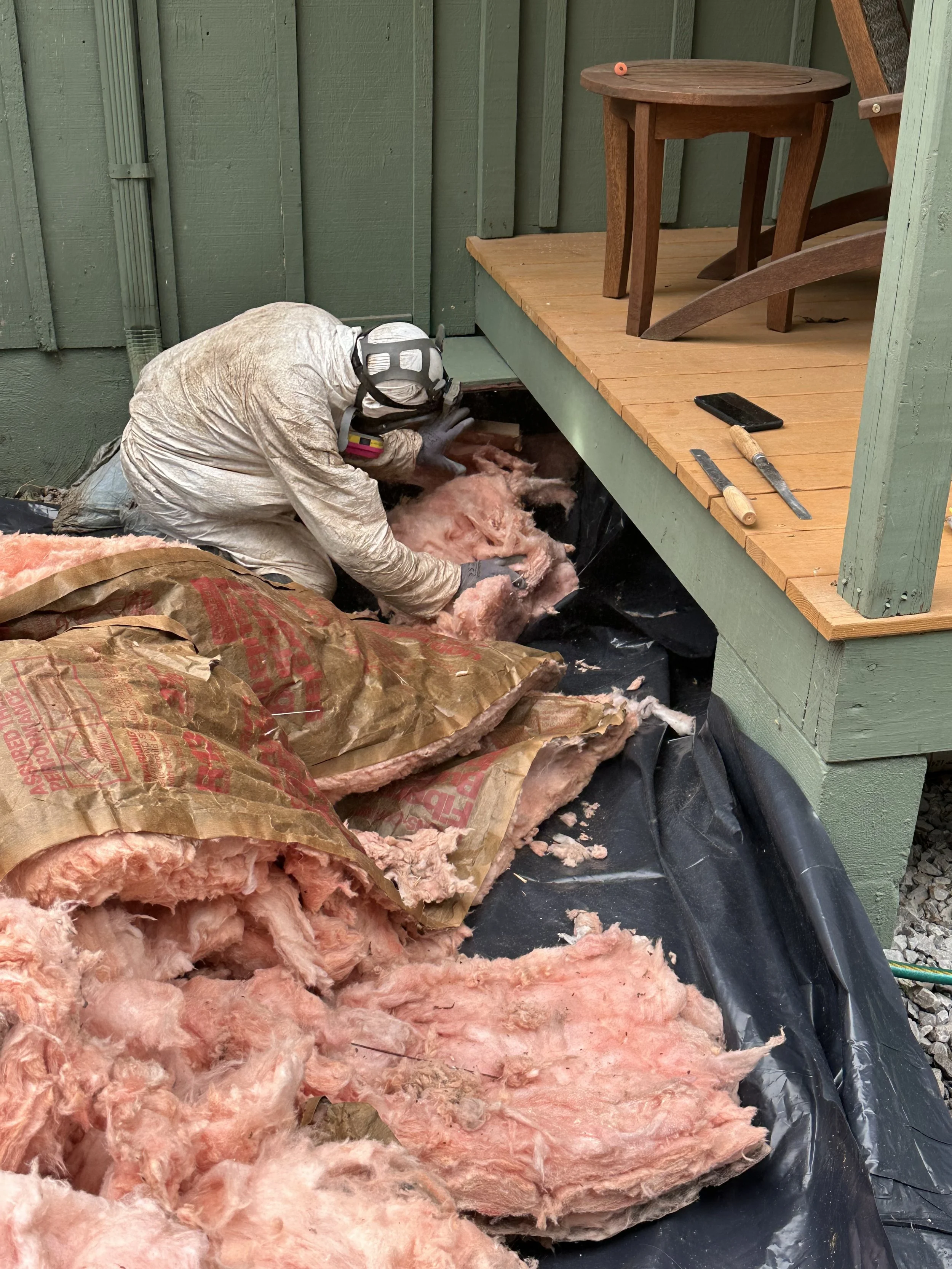 Person wearing protective gear installing insulation in a house wall, with pink fiberglass insulation and tools on a wooden porch.