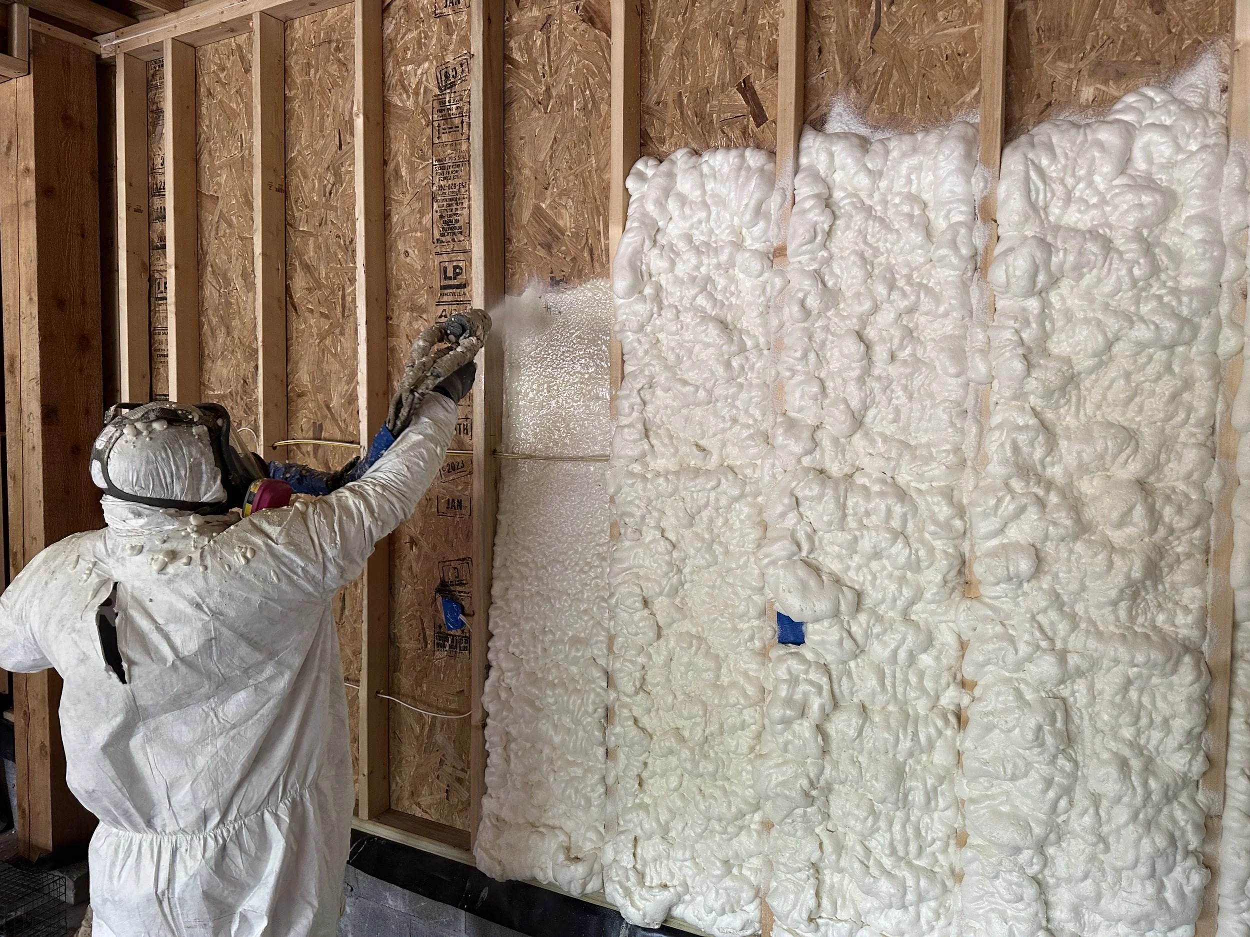 Worker spray foaming wall insulation in unfinished building with exposed wooden studs and OSB sheathing.