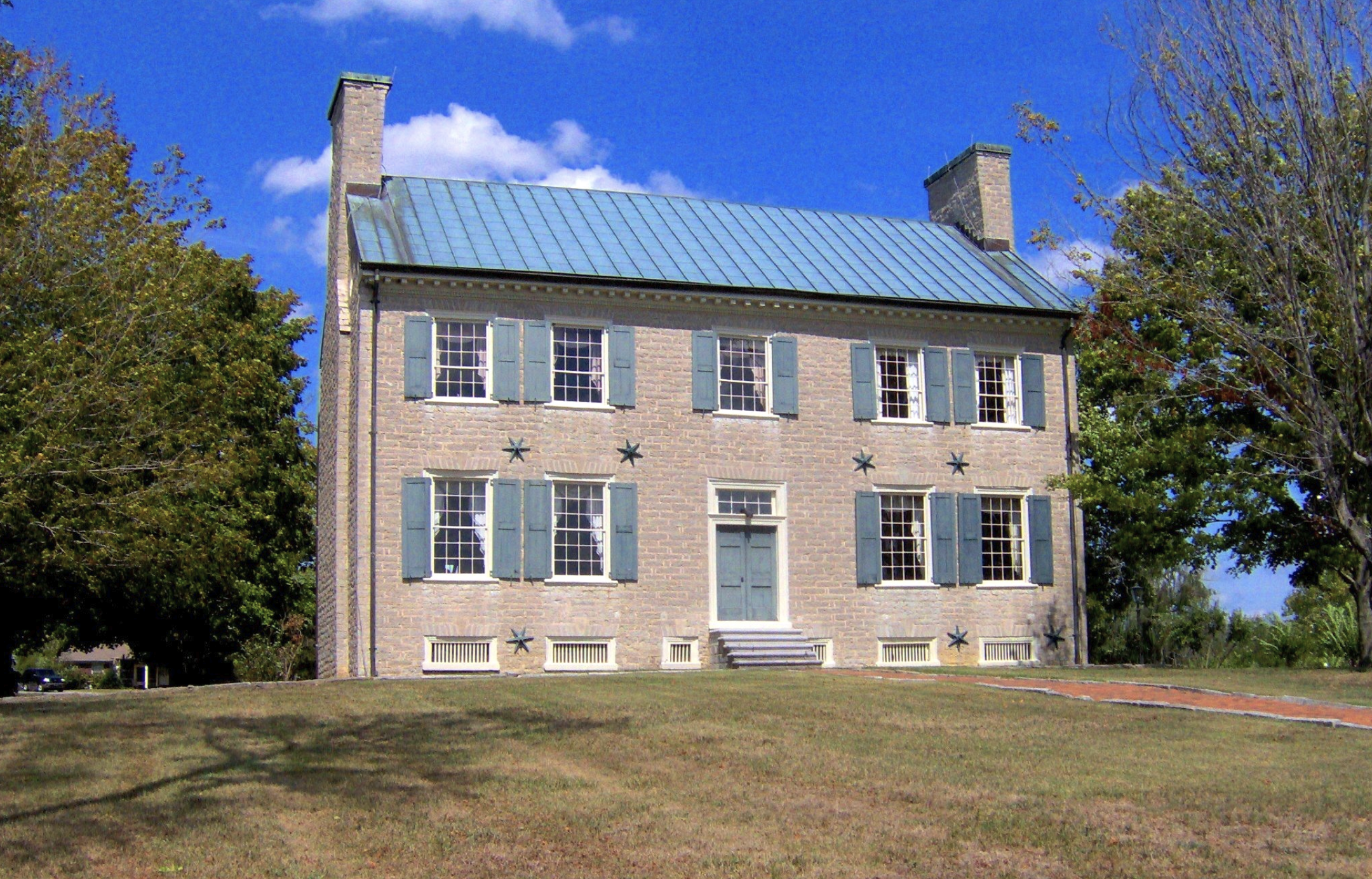 A multi-story brick house with blue shutters and a metal roof, surrounded by trees with green leaves, under a bright blue sky with some clouds.