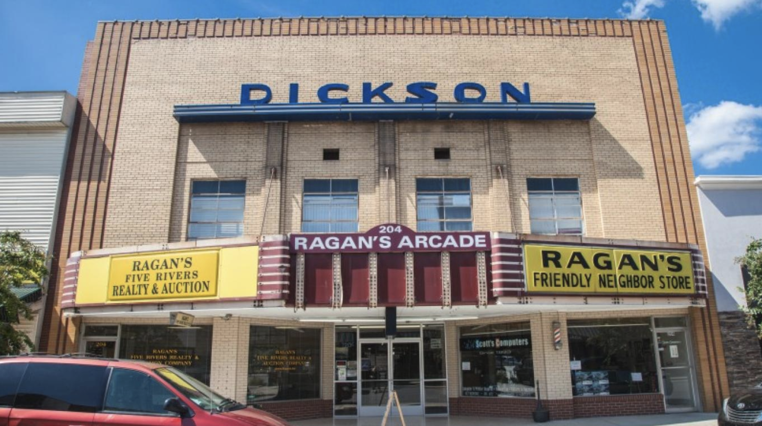 Front view of a building with a sign reading 'PICKSON' on the upper part and a marquee below with signs advertising 'Ragan's Five Rivers Realty & Auction' and 'Ragan's Friendly Neighbor Store,' with parked cars in front and a blue sky with clouds above.