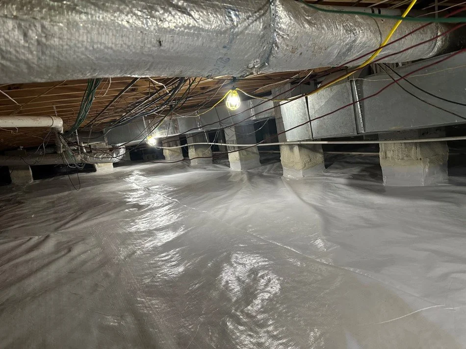 Underground crawl space with plastic vapor barrier on the ground, ductwork, electrical wires, and low-hanging lights.