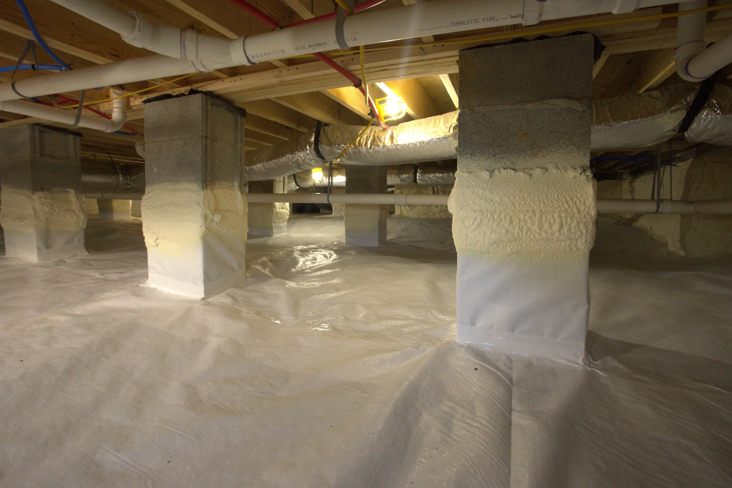 Basement or crawl space with cement block supports, wood framing, PVC pipes, and insulation installed on the floor.