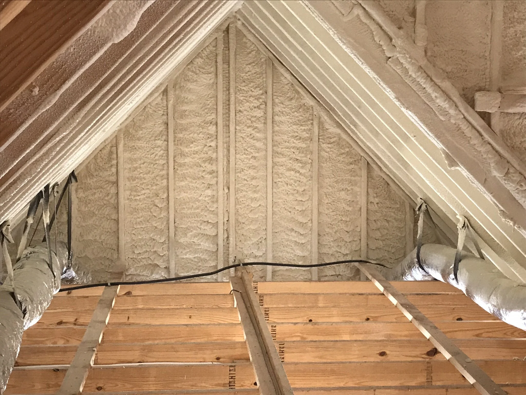 Attic space with wooden beams, insulation spray foam on the walls and ceiling, and ductwork wrapped in insulation.