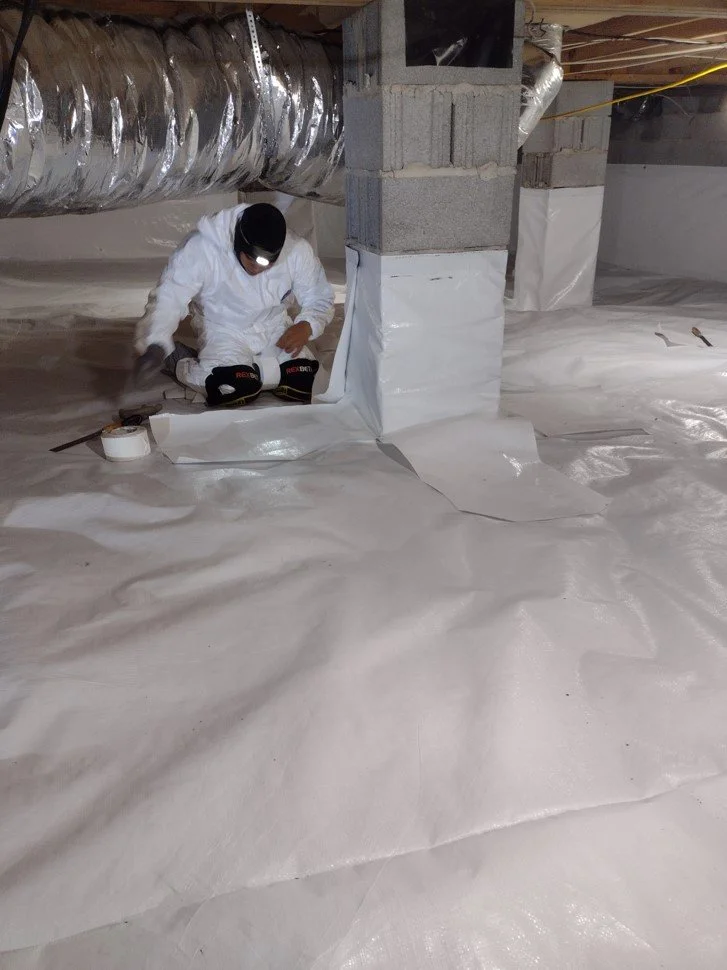 A worker in protective gear installing vapor barrier on a basement floor beneath ductwork and concrete columns.
