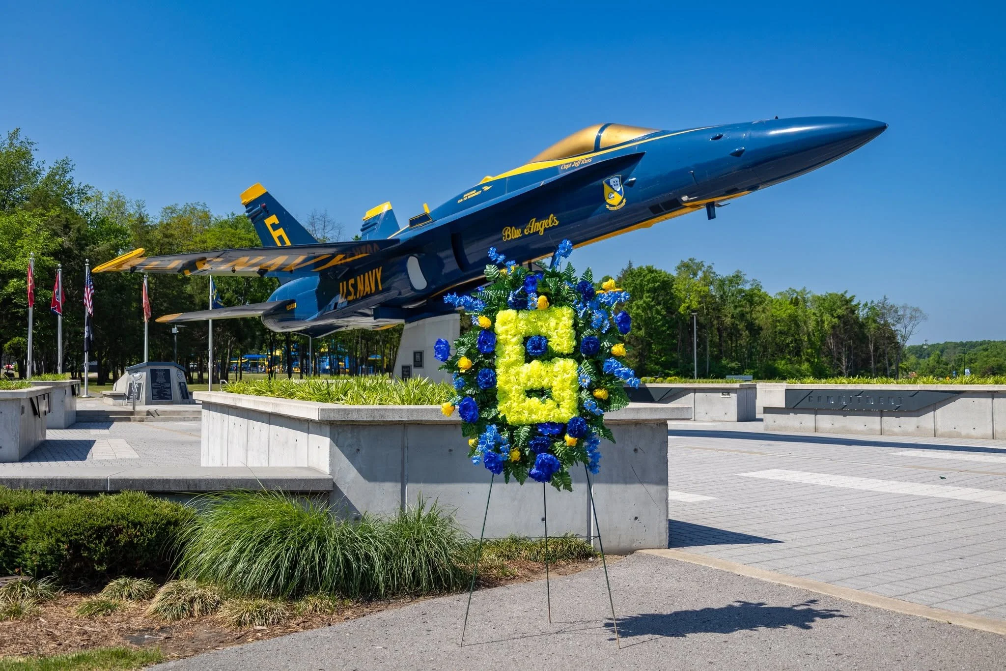 Memorial outdoor display featuring a blue and yellow U.S. Navy fighter jet mounted on a stand, with a colorful floral wreath in front. The background includes trees, flagpoles, and a clear blue sky.
