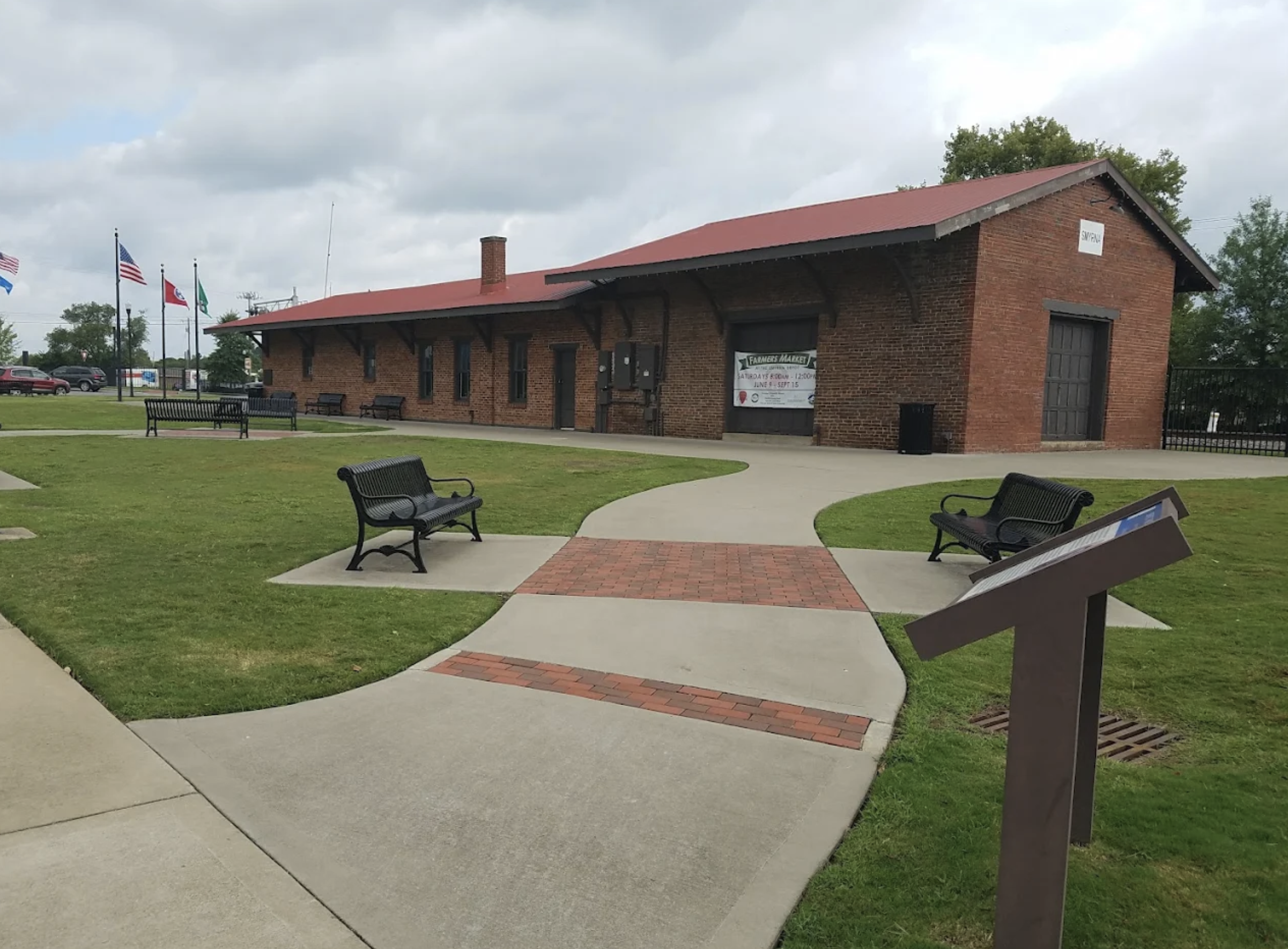 A brick building with a red metal roof, surrounded by a well-maintained grassy area with benches. Flags are flying in the background, and there is a sign on the building indicating a farmers market from 8 am to 12 pm.