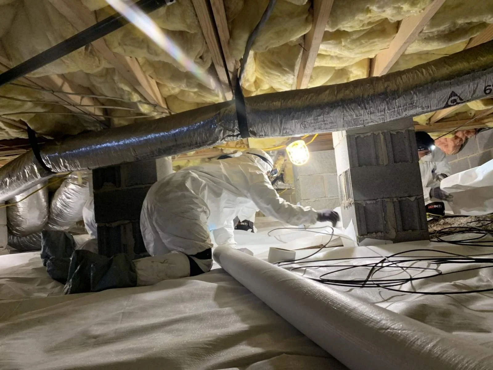 Workers in protective suits installing or inspecting insulation and HVAC ductwork in a basement or crawl space.