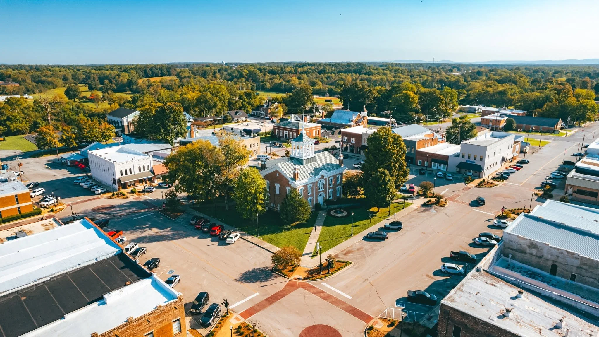 Aerial view of a small downtown area with historic brick buildings, parking lots, and tree-lined streets surrounded by green fields and distant hills on a clear day.