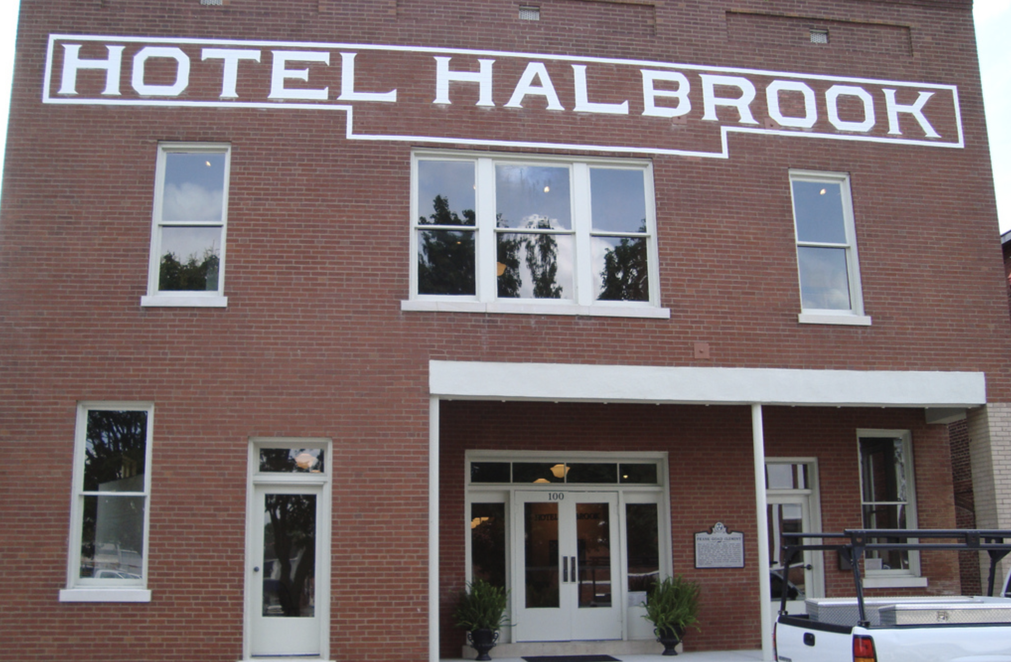 Front of a brick building with a sign that says "Hotel Halbrook" and a street number 100 above the entrance. Reflective windows, plants near the door, and a vehicle in front.