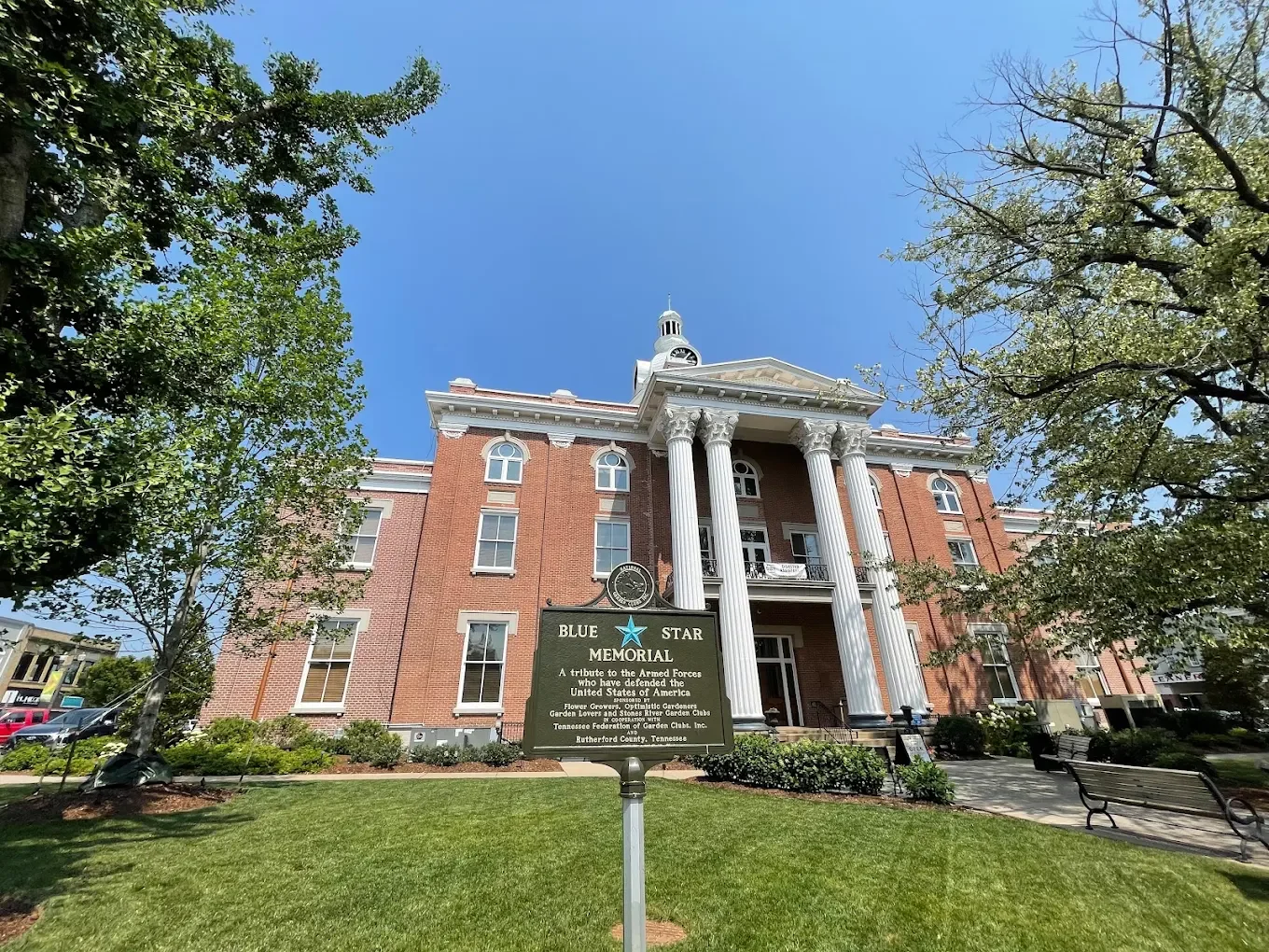 A historic brick building with white columns and a central clock tower, surrounded by green trees and a well-kept lawn, with a sign in the foreground reading 'Blue Star Memorial' honoring the armed forces of the United States.