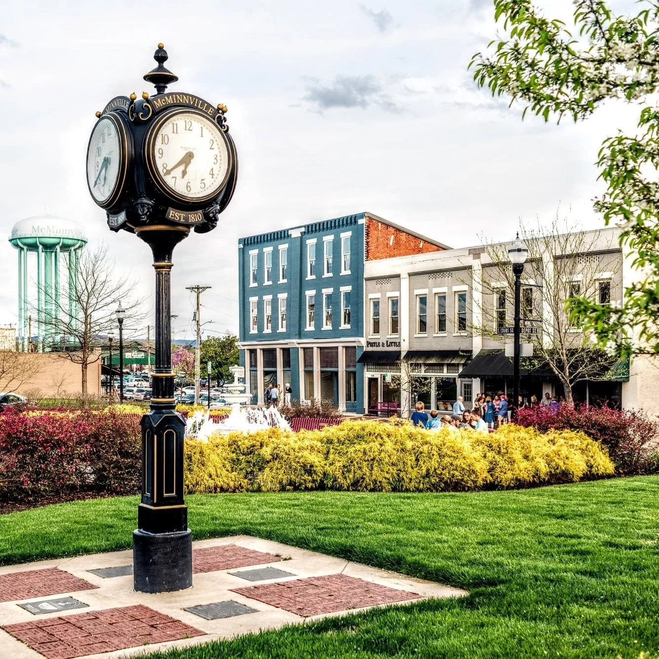 A street scene with a black clock post showing the time as 4:43, surrounded by green grass, colorful bushes, and trees. Several people are walking on the sidewalk in front of multi-story buildings with storefronts, including one named Piola & Luma. The sky is partly cloudy, and there is a water tower visible in the background.