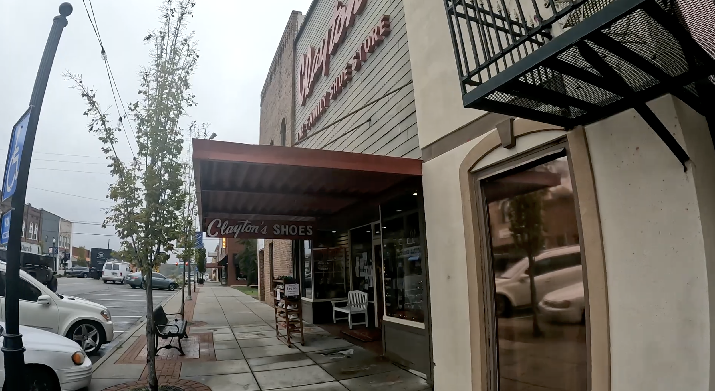 Street view showing storefronts including Clayton's Shoes and a market with signs, parking lot with cars, sidewalk with benches and a tree, overcast sky.