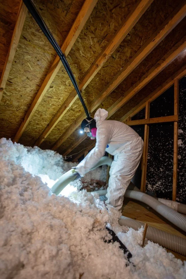 Insulation installer in protective suit working with a hose in an attic with wooden beams and snow insulation on the floor.