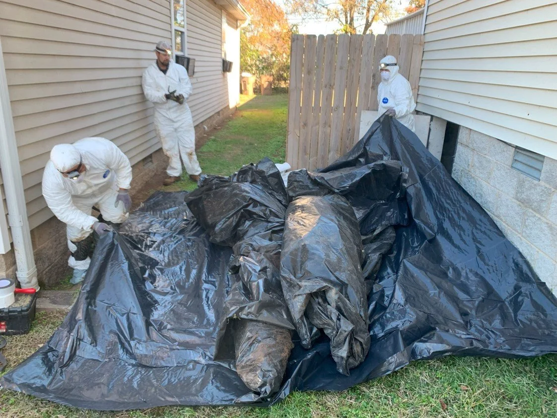 Three individuals dressed in white hazmat suits and protective gear are collecting or managing large black trash bags filled with debris or prohibited materials outside a house, near a fenced yard.