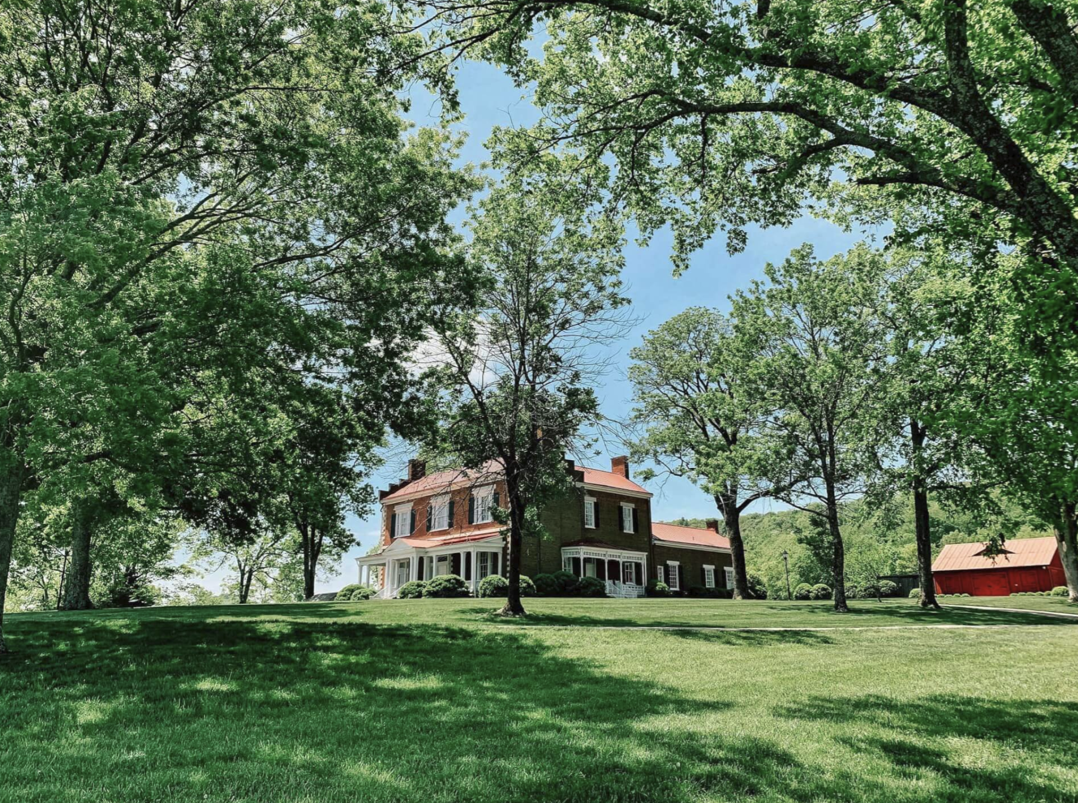 A large, historic, brick house with a wrap-around porch, surrounded by lush green trees and a well-maintained lawn in a suburban neighborhood on a sunny day.