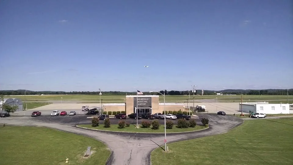 Shelbyville Municipal Airport with parked cars and an airplane flying overhead during the day.