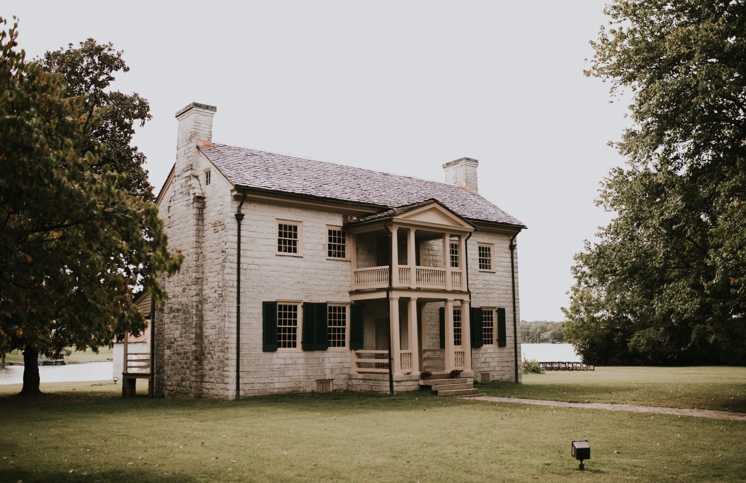A large white brick house with black shutters and a covered front porch sits near a lake, surrounded by green grass and large trees.