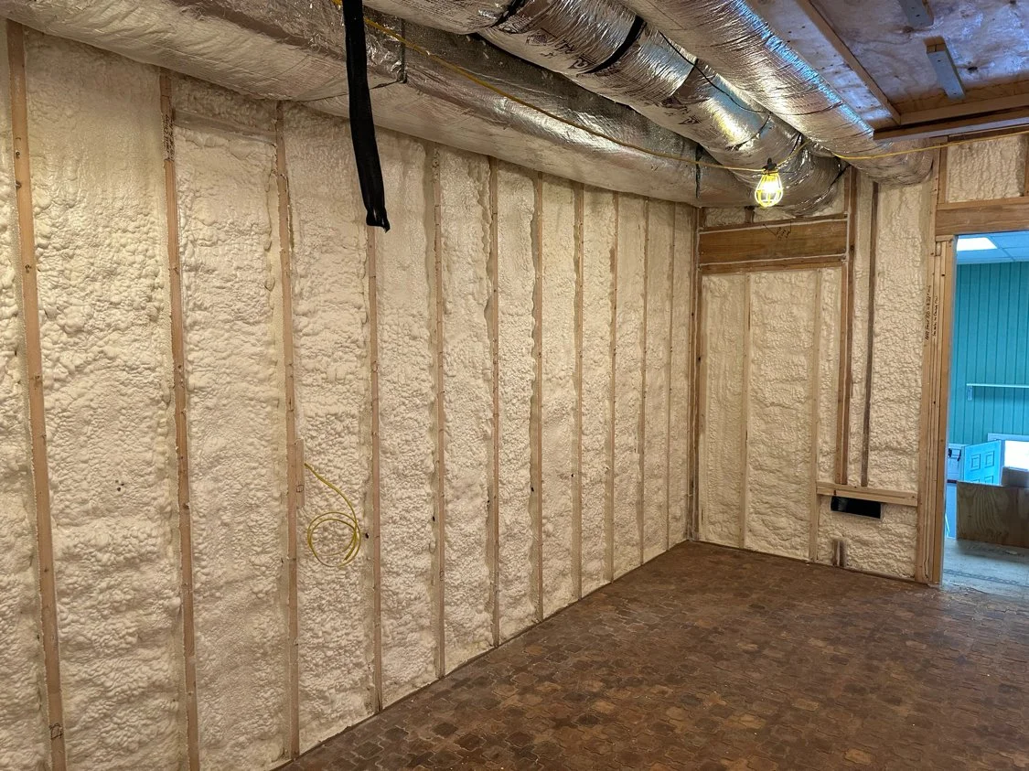 Interior of a room under construction with spray foam insulation on the walls and ceiling, exposed wooden framing, and brick flooring. There is a yellow electrical wire hanging from the wall and a temporary light fixture hanging from the ceiling.