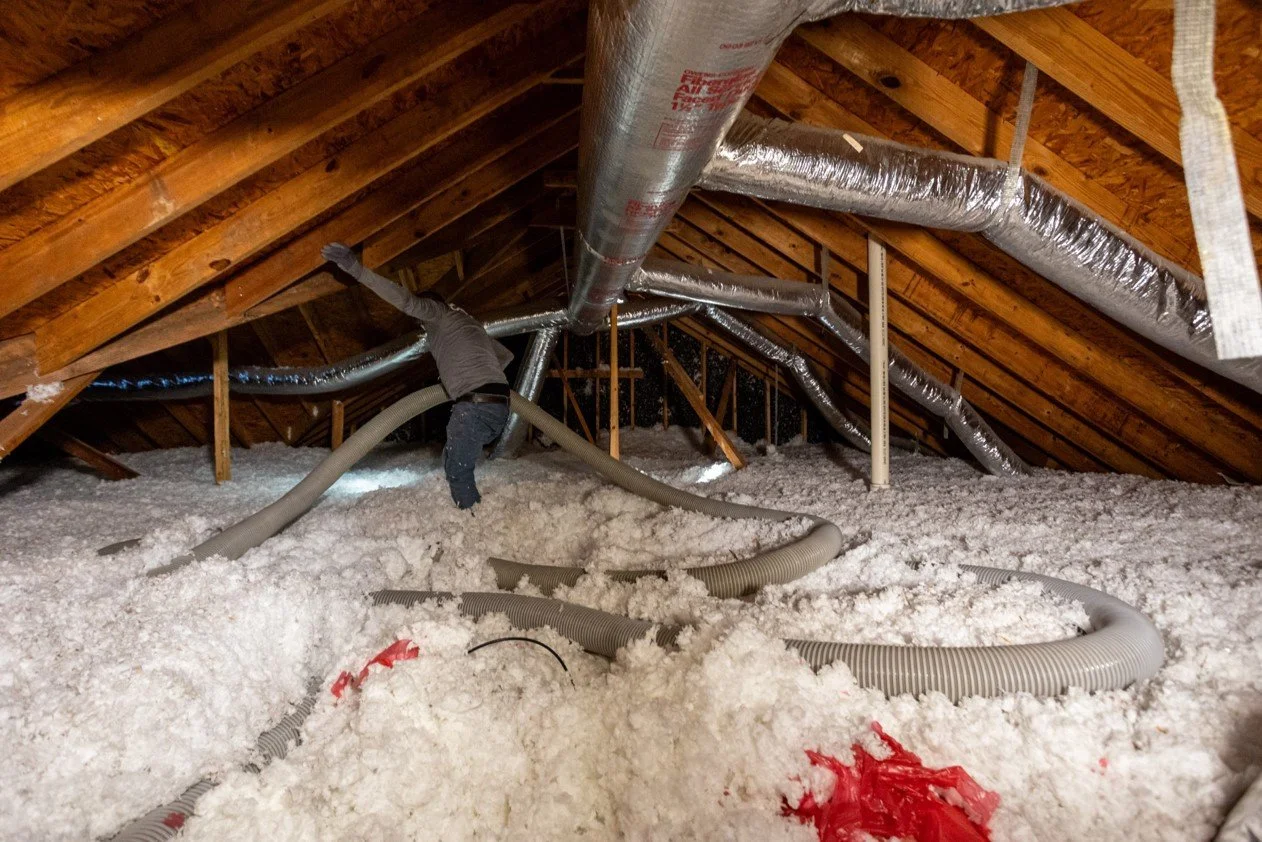 A person working inside an attic with insulation on the floor, using flexible hoses connected to a ventilation system, surrounded by wooden beams and ducts.