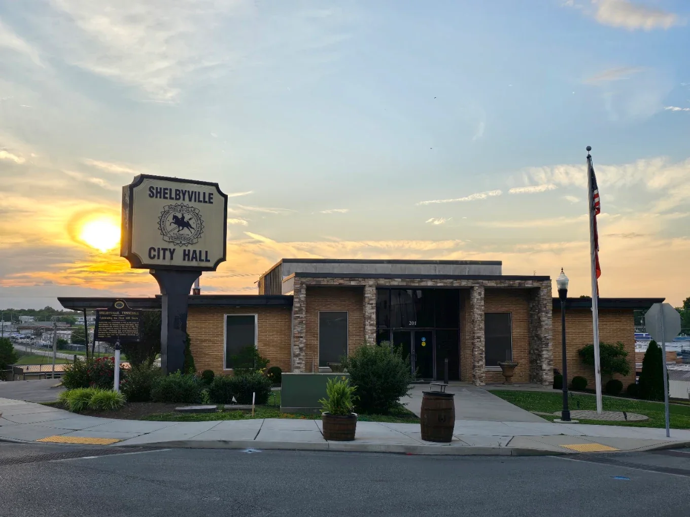 Front view of Shelbyville City Hall building during sunset, with a sign reading 'Shelbyville City Hall', a flagpole with the American flag, and landscaped plants and sidewalk in the foreground.