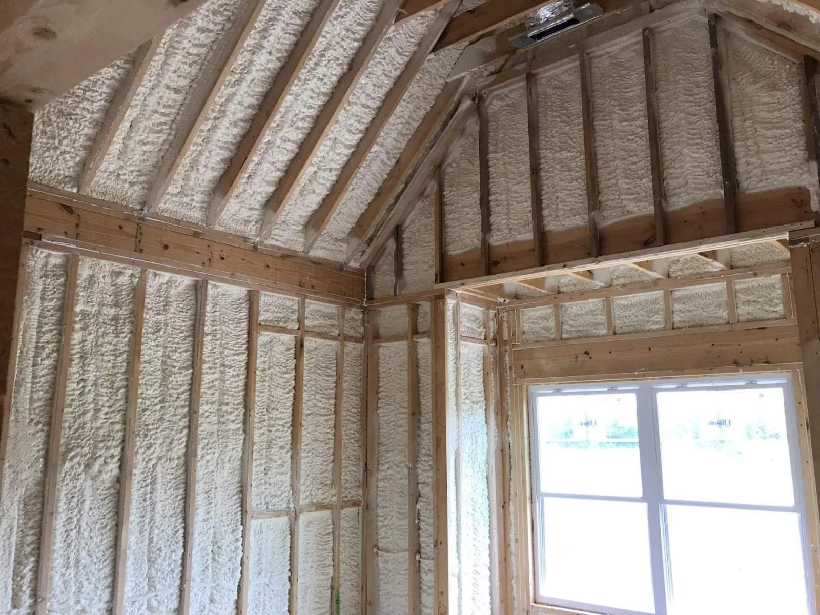 Interior of a room under construction with foam insulation sprayed on the walls and ceiling, exposed wooden framing, and a large window.