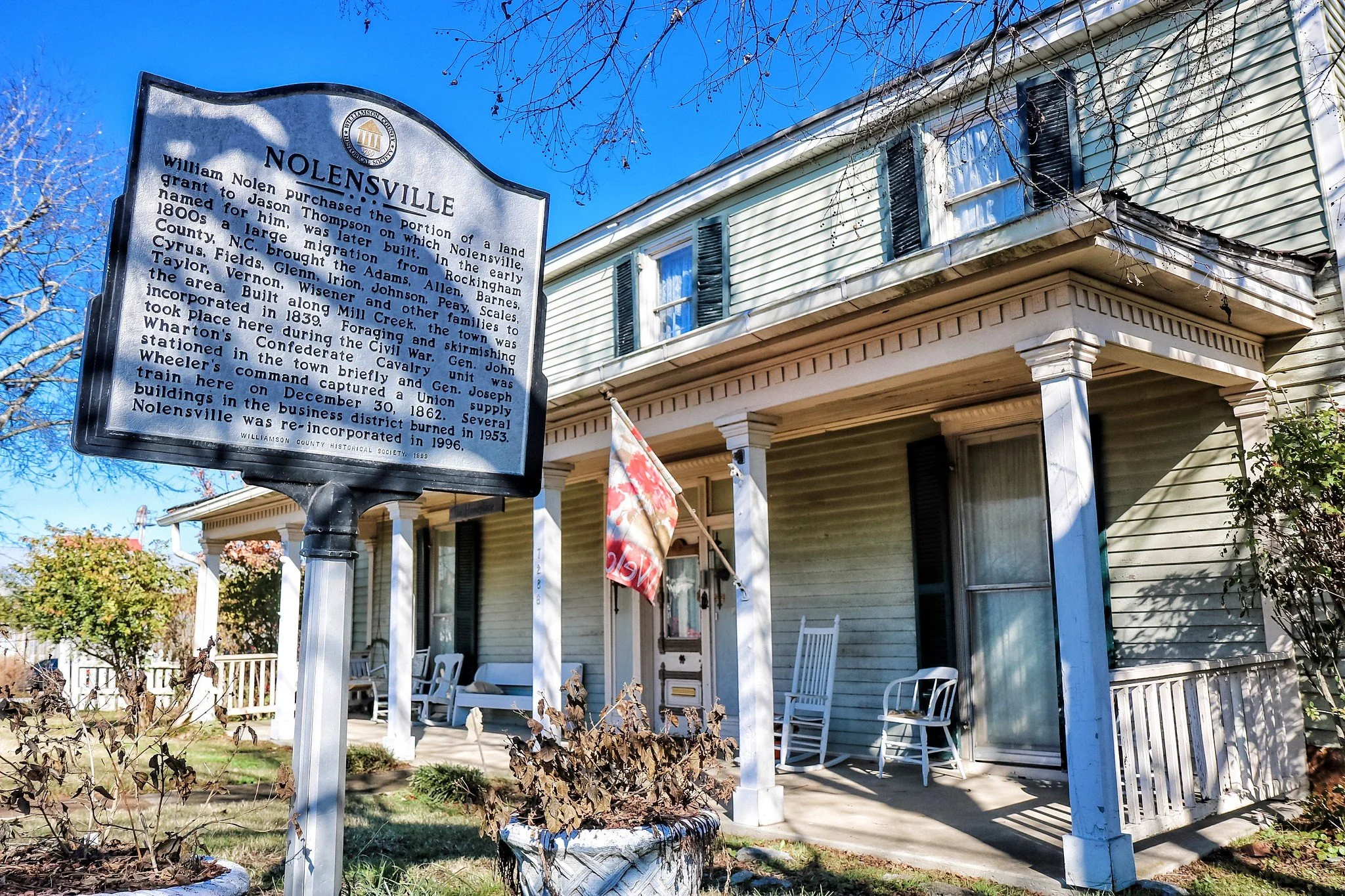 Historical marker for Nolen'sville in front of a house with porch in a suburban neighborhood. The house has light green siding, black shutters, and white columns. There are trees with bare branches and a blue sky.