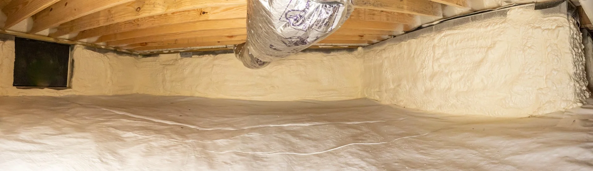 Insulation foam sprayed on crawl space, with wooden beam ceiling and plastic vapor barrier.