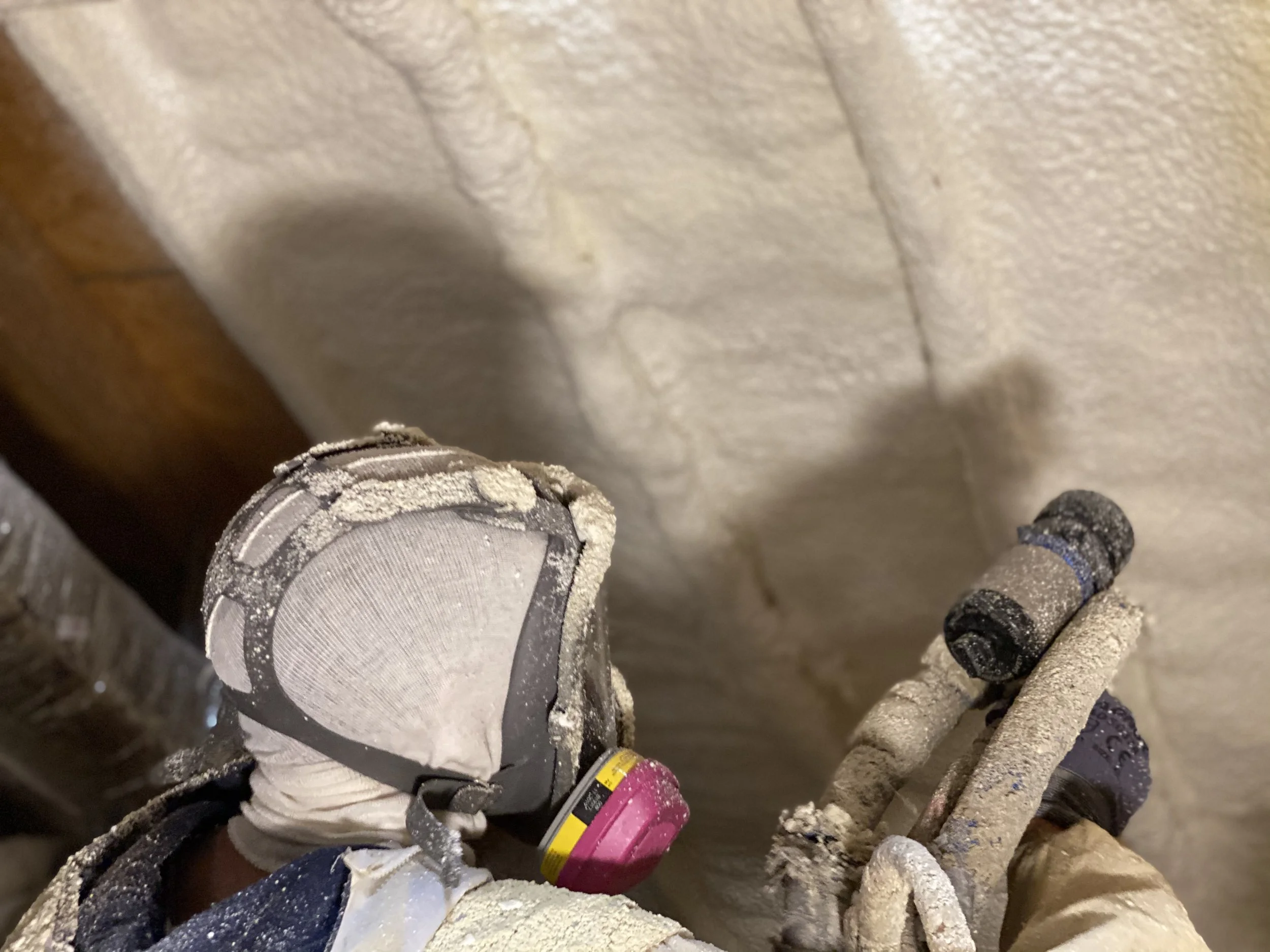 Worker wearing protective gear spraying foam insulation on attic ceiling.