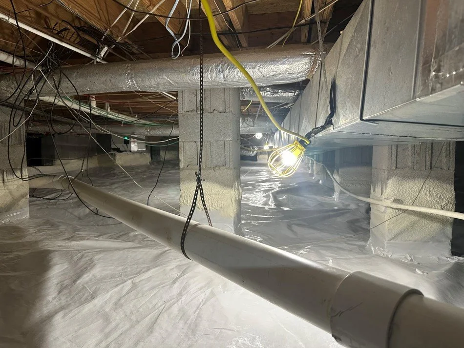Underground construction site with exposed pipes, wiring, support beams, insulation, and a portable work light hanging from the ceiling.