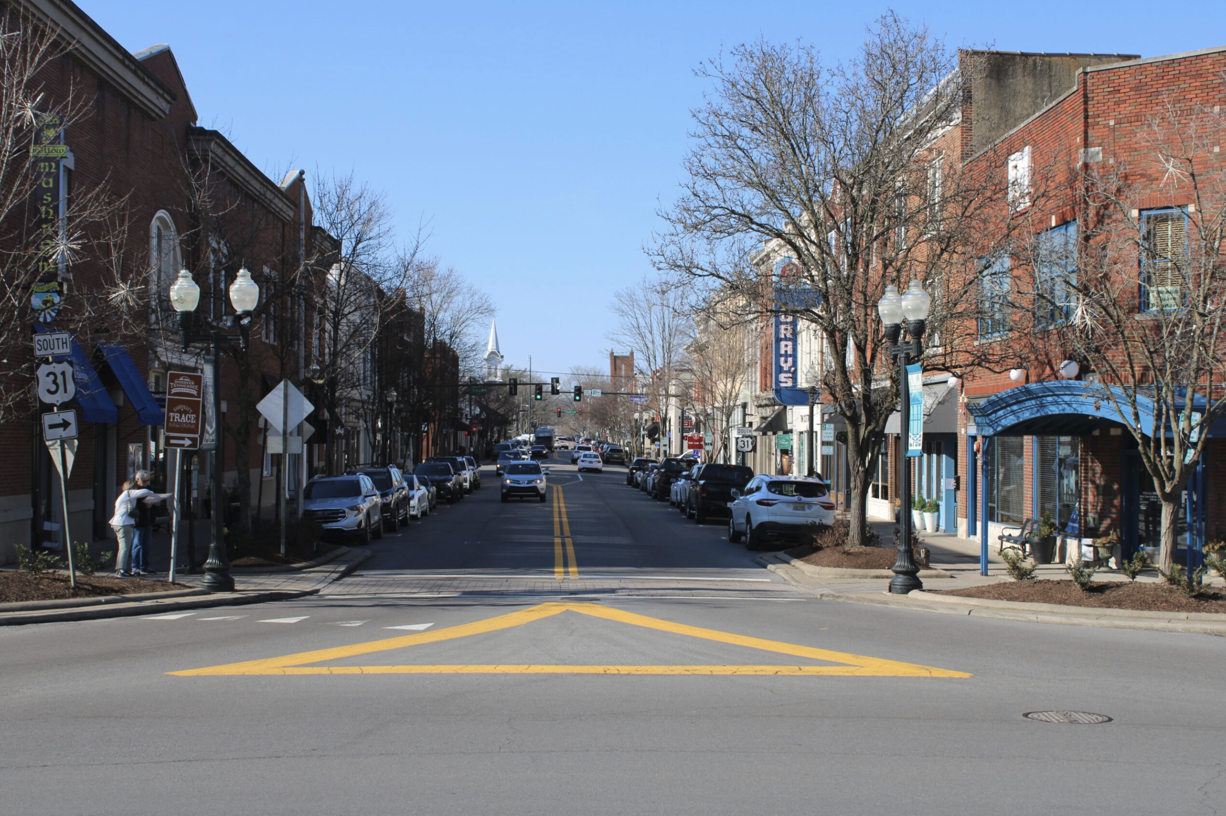 A downtown street scene with parked cars on the sides, leafless trees, brick buildings, and a church steeple in the background under a clear blue sky.