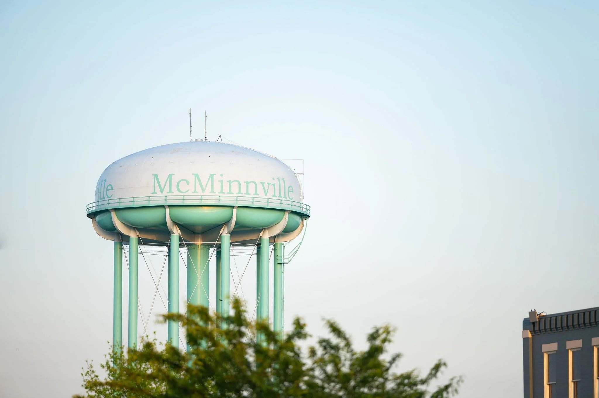 The McMinnville water tower with a rounded top and support beams, visible against a clear sky, partially obscured by trees in the foreground.