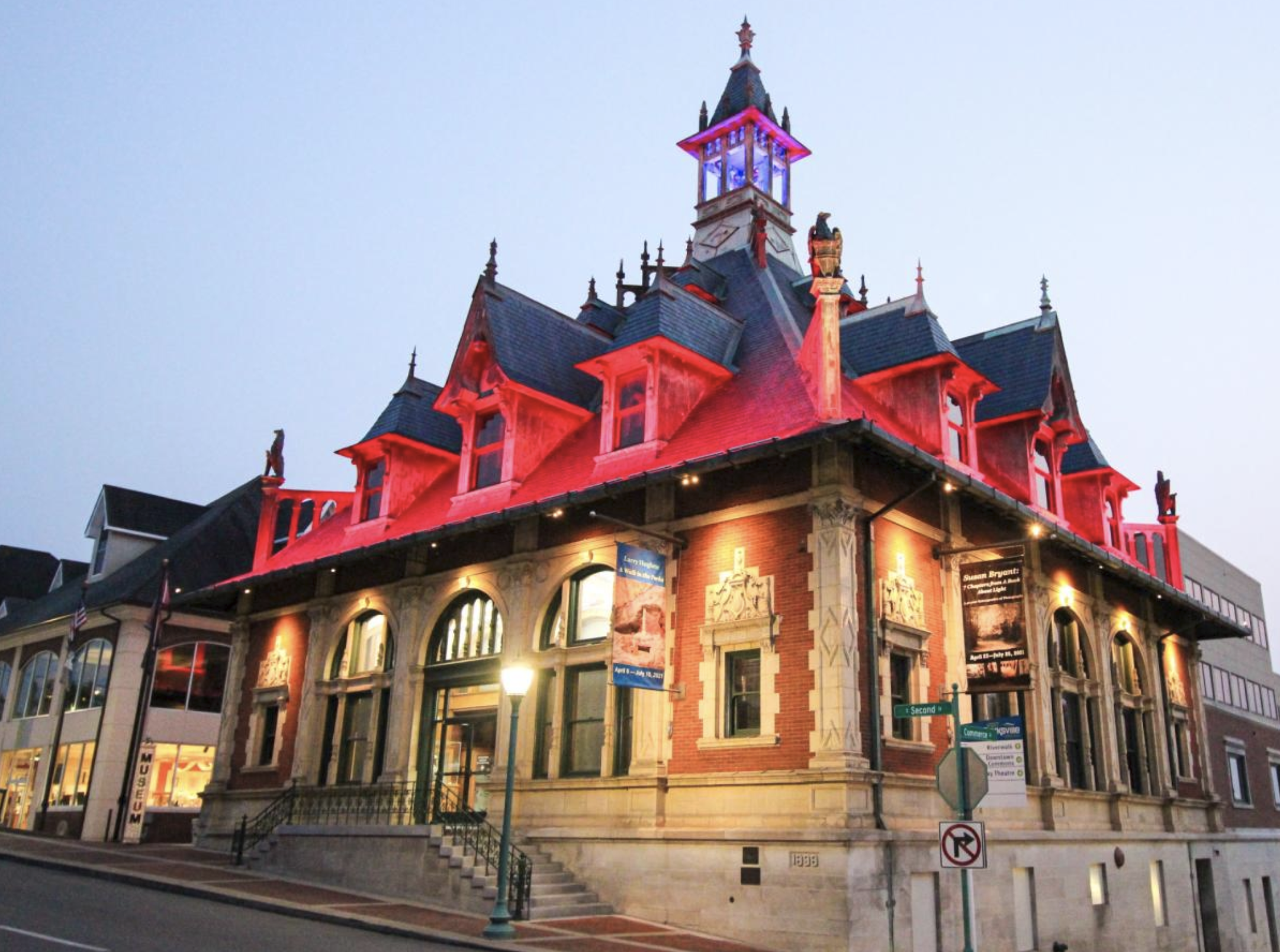 A historic building decorated with red lighting at night. The building features ornate architectural details, including a tall spire with a clock and decorative statues on the roof. Street signs and a lamppost are visible in front of the building.