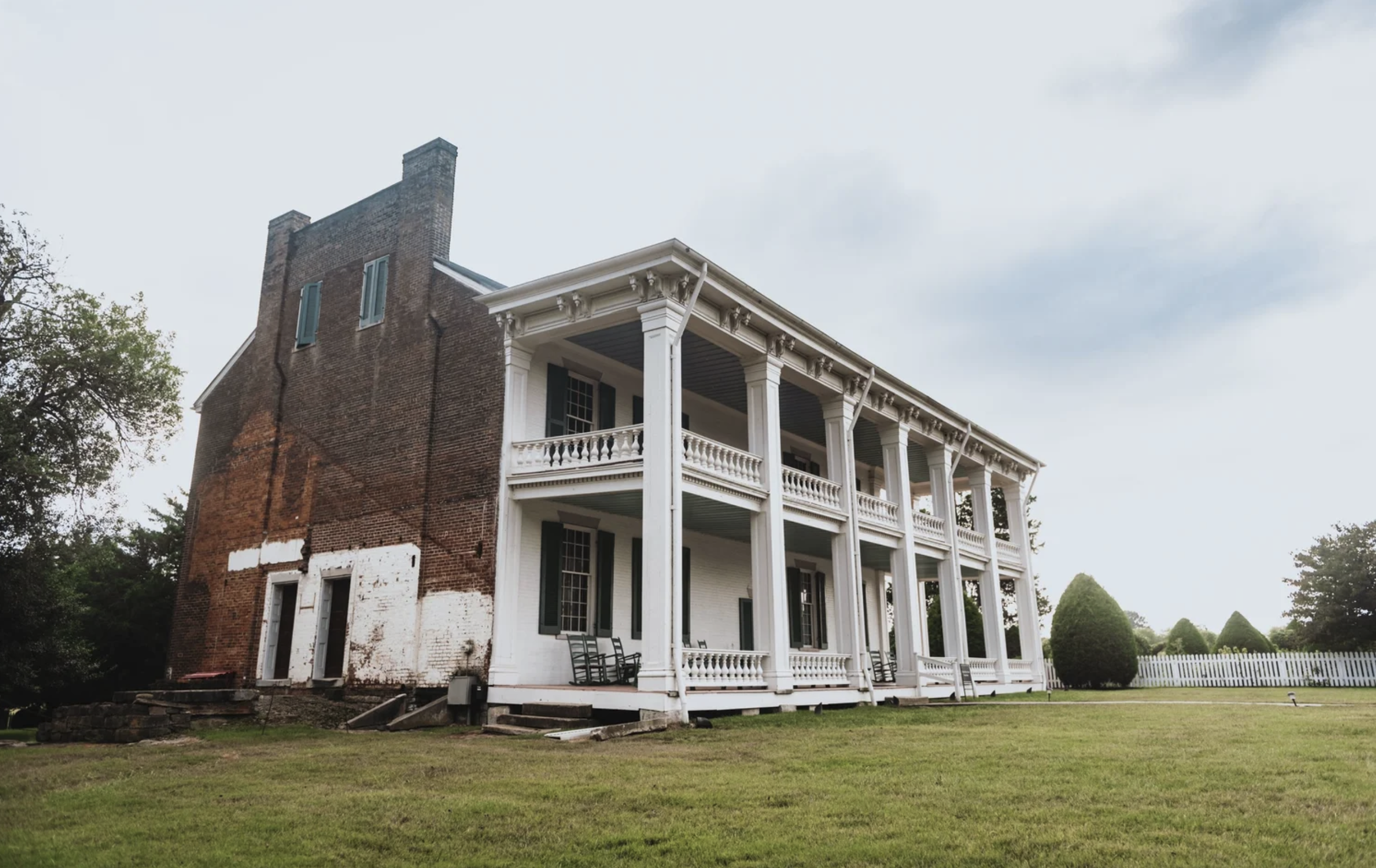 A large historic brick house with white columns and trim, multiple balconies, and rocking chairs on the porch, surrounded by a well-maintained lawn and trees.