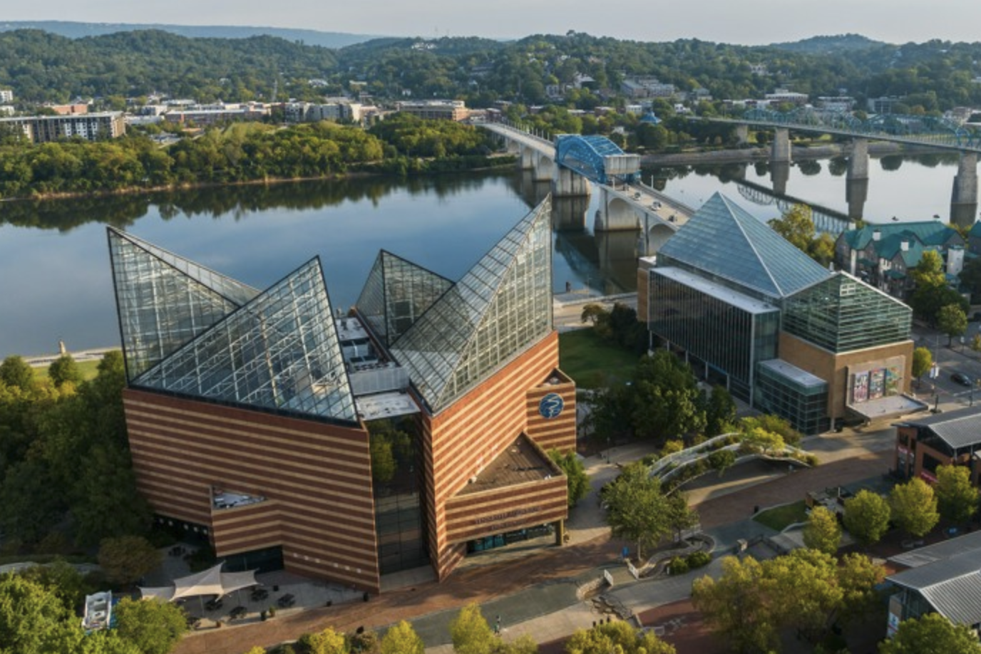 Aerial view of a modern cityscape with a river, a bridge, and several contemporary buildings, including a distinctive structure with a glass pyramid roof, green trees lining the streets, and hills in the background.