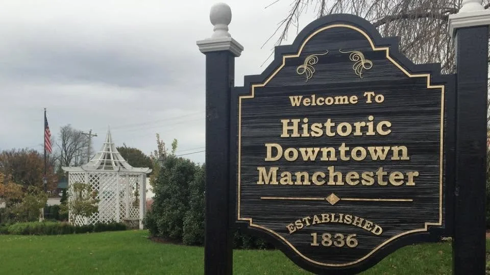 A sign welcoming visitors to Historic Downtown Manchester, established in 1836, with a gazebo and American flag in the background.