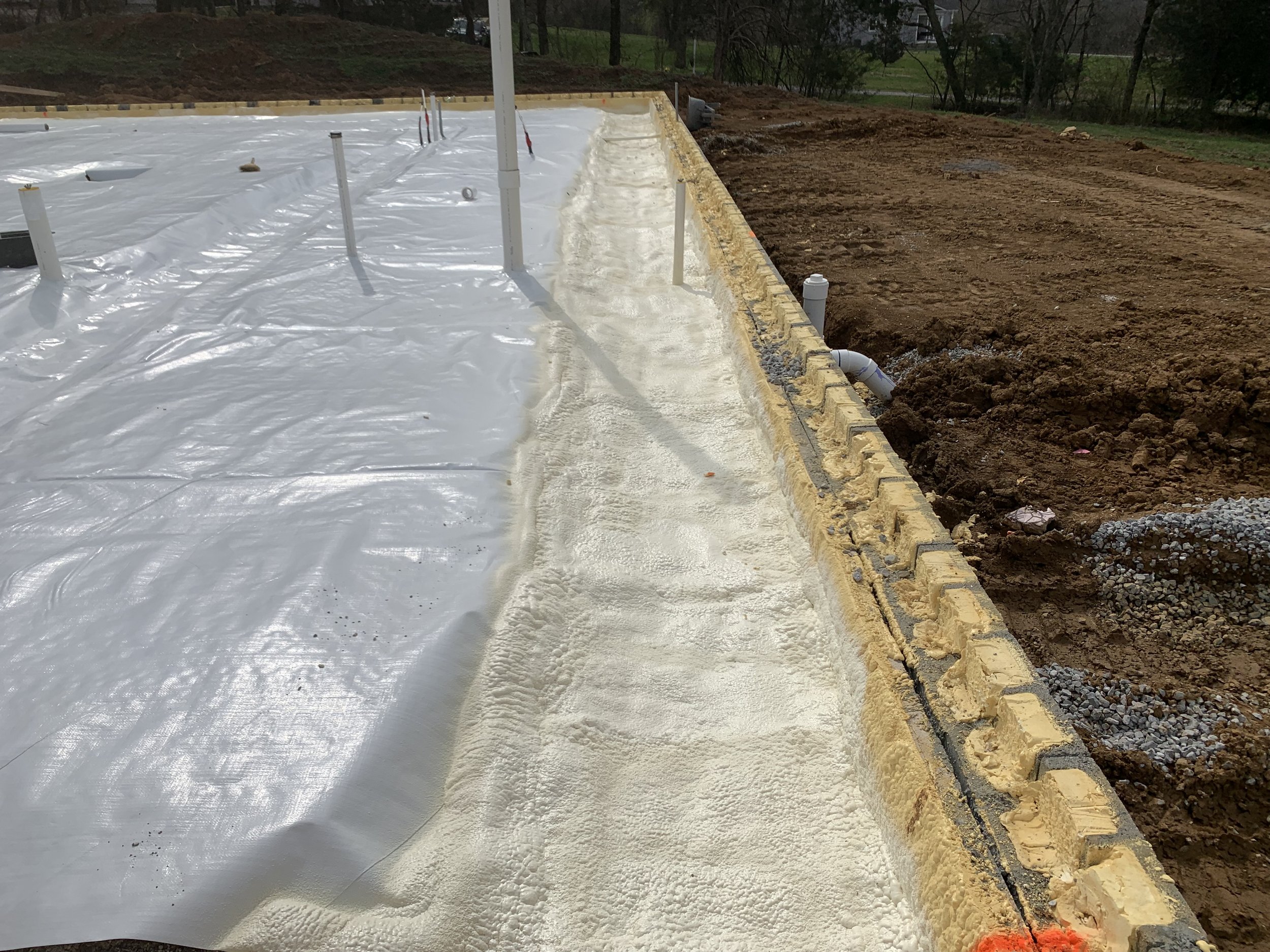 Construction site with a concrete foundation covered by a plastic sheet and spray foam on foundation.